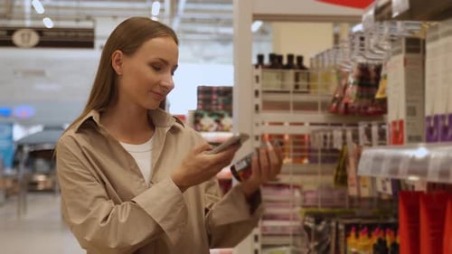 A Woman in a Beige Shirt Scans a Package of Cosmetics in a Retail Store She is Using a Smartphone to
