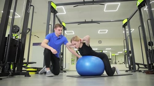 Woman Exercises on Ball with Trainer in Gym
