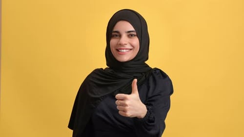 Lächelnde junge saudische Frau in schwarzem Hijab, gestikuliert coolen Daumen hoch, gute Wahl, isoliert, orange