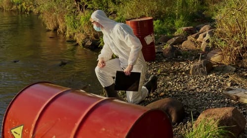 Un travailleur environnemental en équipement dangereux examine un baril rouge au bord du lac tout en évaluant la possibilité