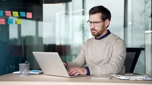 Frustrated businessman struggling with laptop while sitting at desk in office.