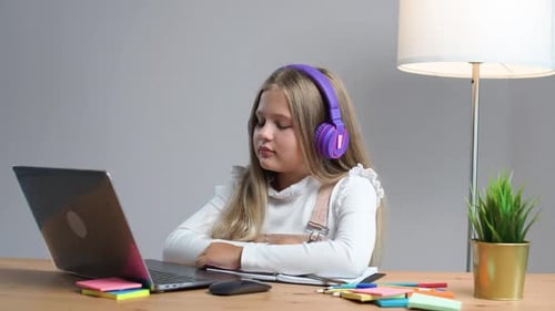 Distance Learning School Girl in a Purple Headphones Sitting at the Table at Home and Uses a Laptop