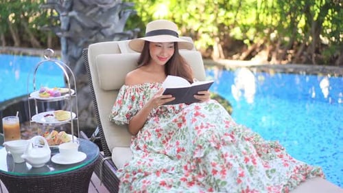 Young beautiful lady wearing a dress and sun hat, relaxing near a swimming pool reading a book and t