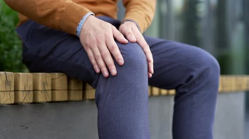 Male hands massaging knee leg muscles while sitting outside on a bench on a city street. Businessman