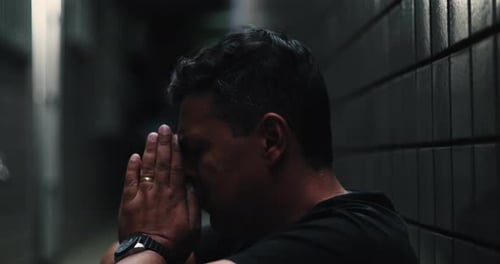 Man sitting against a wall in a narrow alley, head tilted upward, facial expression of deep pain