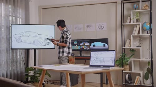 Asian Male Comparing His Car Design On A Tablet To The Photo On Tv While Working In The Studio