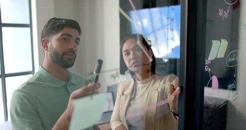 Discussing project strategy, colleagues writing on glass board in modern office