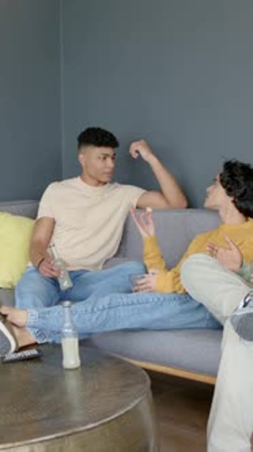 Vertical video: Relaxing men starting chat on sofa at home when left man lifting bottle eating chip