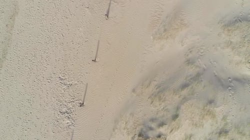 Long fence post shadows stretch out across a smooth sandy beach covered in footprints, aerial god vi
