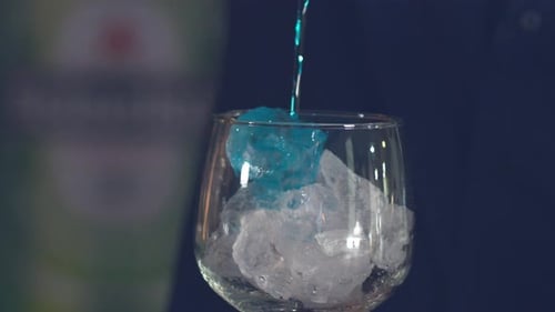 Slow motion, pouring blue liquor from bottle into cocktail glass with ice. Close Up