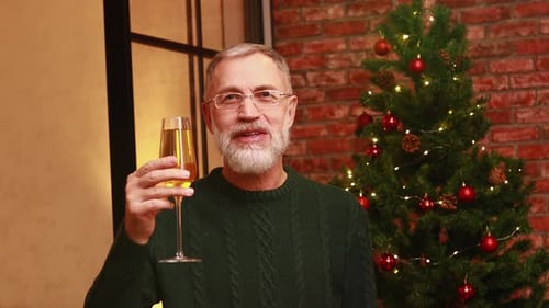 an Elderly Man in a Green Knitted Sweater Raising a Toast with Champagne Near the Christmas Tree