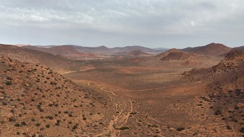 Aerial ascends from red, dry, arid, hilly desert landscape: cloud sky