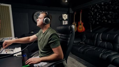 a focused male sound engineer wearing headphones sits at a mixing desk recording new track