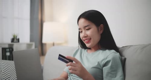 Young woman uses laptop and credit card to shop online on sofa at home.