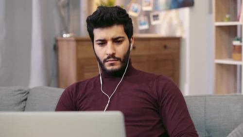 Rotation View of a Young Bearded Man in Headphones Actively Using His Laptop Being Online Gaming