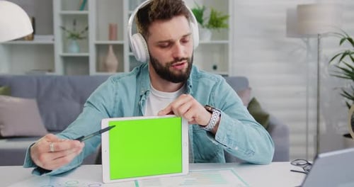 Hermoso joven sonriente que sostiene una tableta de pantalla verde mientras chatea en línea con sus socios. Varón