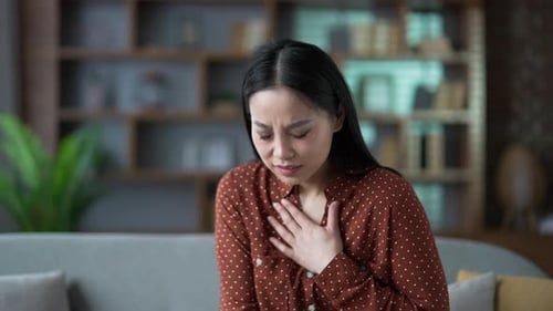 Young asian woman has a heart attack while sitting on sofa in the living room at home. Sick female