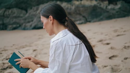 Junge Frau liest Buch und sitzt am Sandstrand in weißem Hemd, Nahaufnahme