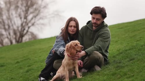 A Young Couple with Their Dog Sits on the Grass in the Park A Bearded Man and His Woman are Sitting