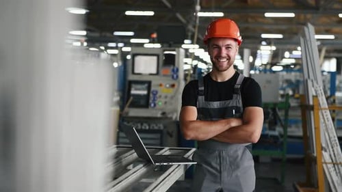 Smiling positive worker in grey uniform stands indoors in the factory with arms crossed