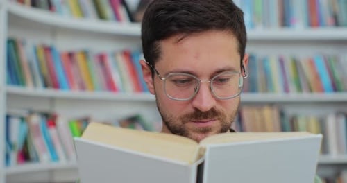 Portrait of Man Student in Glasses is Spending His Time in Library Examining a Book and Reading the