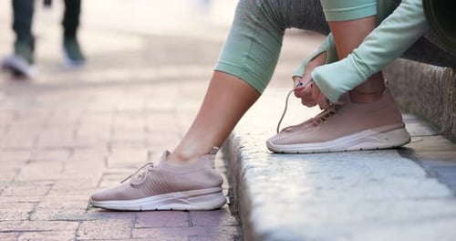 Runner sitting on stone curb is tying pale pink sneaker laces by walkway, preparing to run