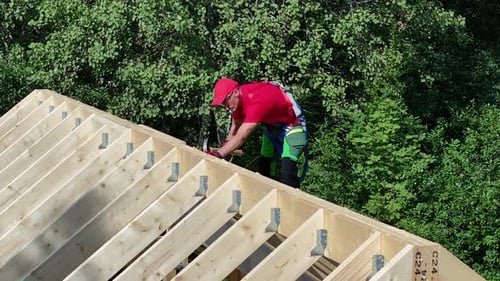 Construction worker installs roof trusses in green wooded area during sunny day