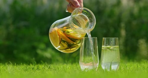 Hand Pouring Refreshing Lemonade with Mint and Lemon Slices Into Glasses on a Lush Green Lawn