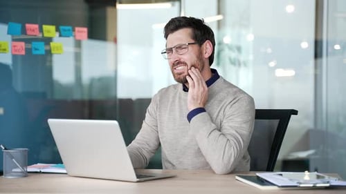 Businessman suffering from toothache while sitting at desk in modern office. Mature professional