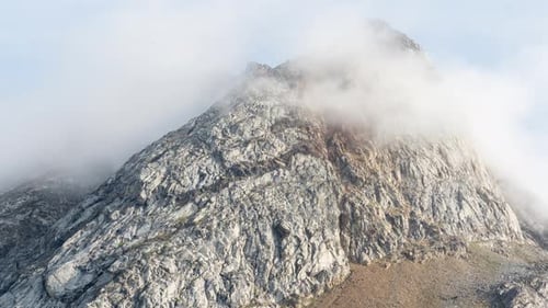Fog and clouds move over a rocky cliff face and mountain peak. Time lapse view of weather change on