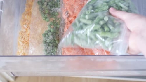 Female hand holds a plastic bag with frozen green beans on the background of freezer.
