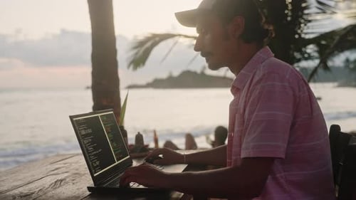 Desenvolvedor de software trabalha em laptop no Beachside Cafe Man escreve código durante o pôr do sol no oceano, palmeiras balançam