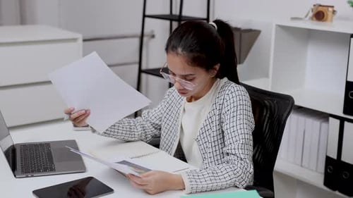 asian woman focusing paperwork checking notes and typing laptop sitting at bright home office desk
