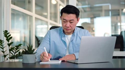 Man Working At Desk Writing and Using Laptop