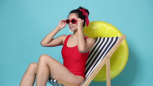 Mujer con traje de baño y gafas de sol en tumbona sobre fondo azul claro