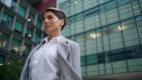 Confident female executive strolling downtown looks at camera near glass office building