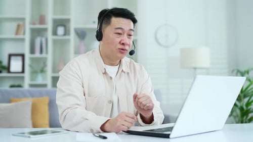 Confident asian businessman in headset talking on video call on laptop while sitting in home office.