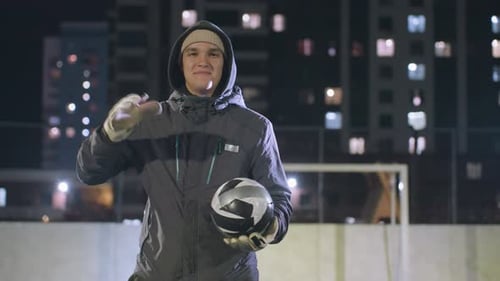Smiling Man in Hoodie Hitting Soccer Ball on Urban Field at Night