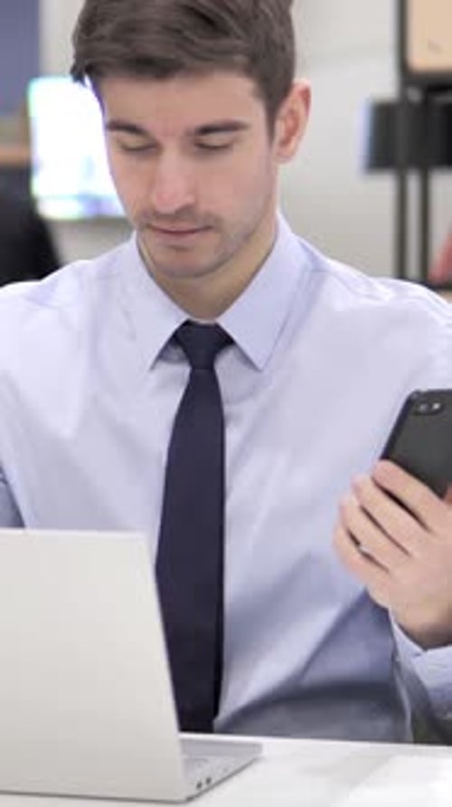 Young Adult Using Smartphone at Desk with Laptop