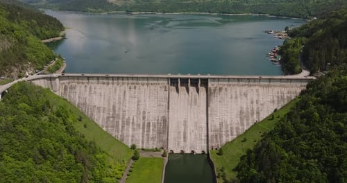 Dam and water reservoir surrounded by green forests, panoramic aerial view. Tracking drone shot, sun