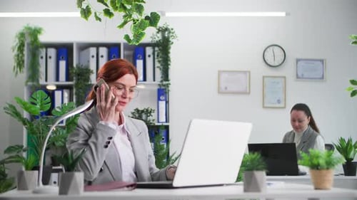 Jeune femme souriante à lunettes parlant au téléphone tout en travaillant au bureau assise au bureau d'ordinateur