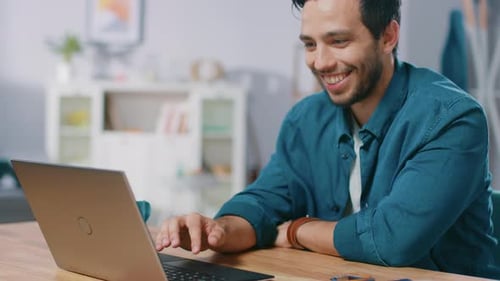 Handsome Young Man Works on a Laptop while Sitting at His Desk and Drinking Beverage in His Living
