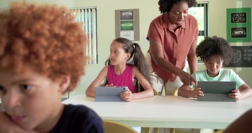 Teacher with students, noticing screens, leaning and adjusting tablet, guiding learning in class