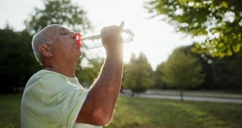 Senior Retired Grey Haired Man Drinking a Bottle of Water Outdoors in a Park After Exercising in a