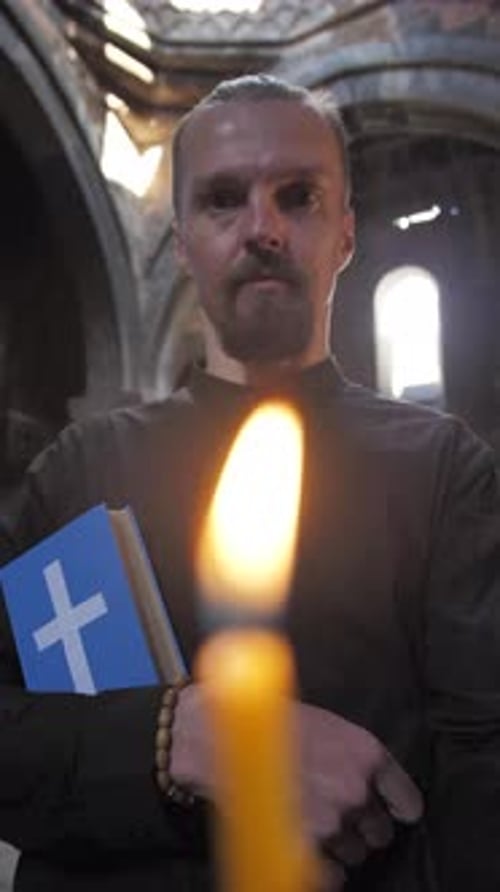 A vertical portrait of a young Caucasian priest holding out a lit church candle in his hand and look