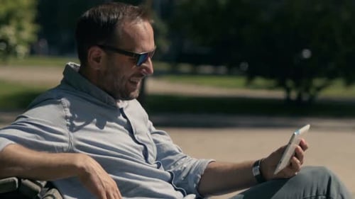 Man Watching Movie On Tablet Computer Sitting On Bench In City Park