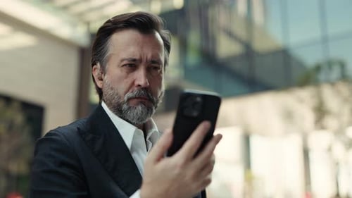 Portrait of Mature Businessman Looks at Smartphone Against Business Center