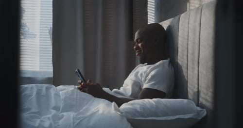 Un homme heureux s'allonge confortablement dans une chambre moderne à l'aide d'un smartphone tout en s'appuyant sur des oreillers moelleux
