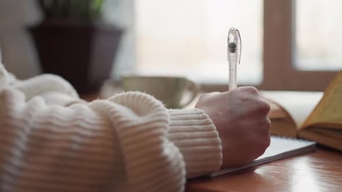 Close Up Female Hand Writing with Transparent Pen Beside Open Book on Desk