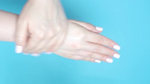 Female hand applies and rubs moisturizer on blue background, top view.